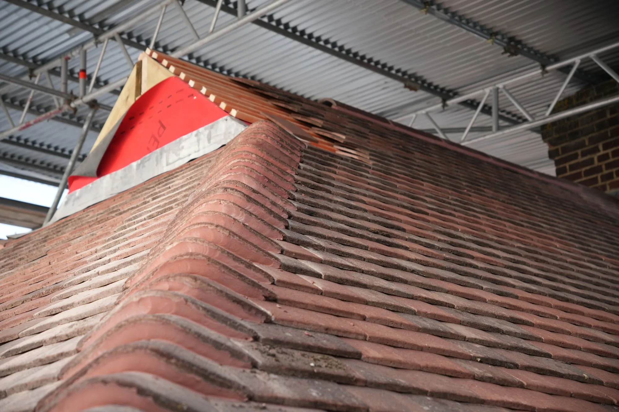 The ridge of the redesigned Brentwood roof mid tiling, membrane and battens still exposed above the new plain tiles, under the scaffold's temporary roof.