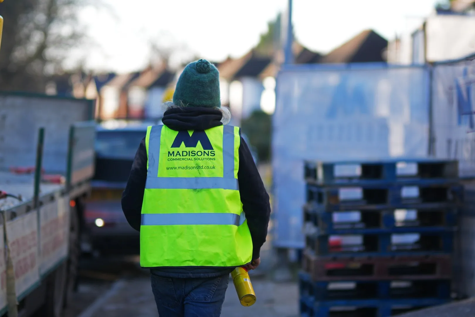 An operative in a Madisons branded hi-vis vest, photographed from behind, walking up the site access past stacked pallets and the delivery lorry.