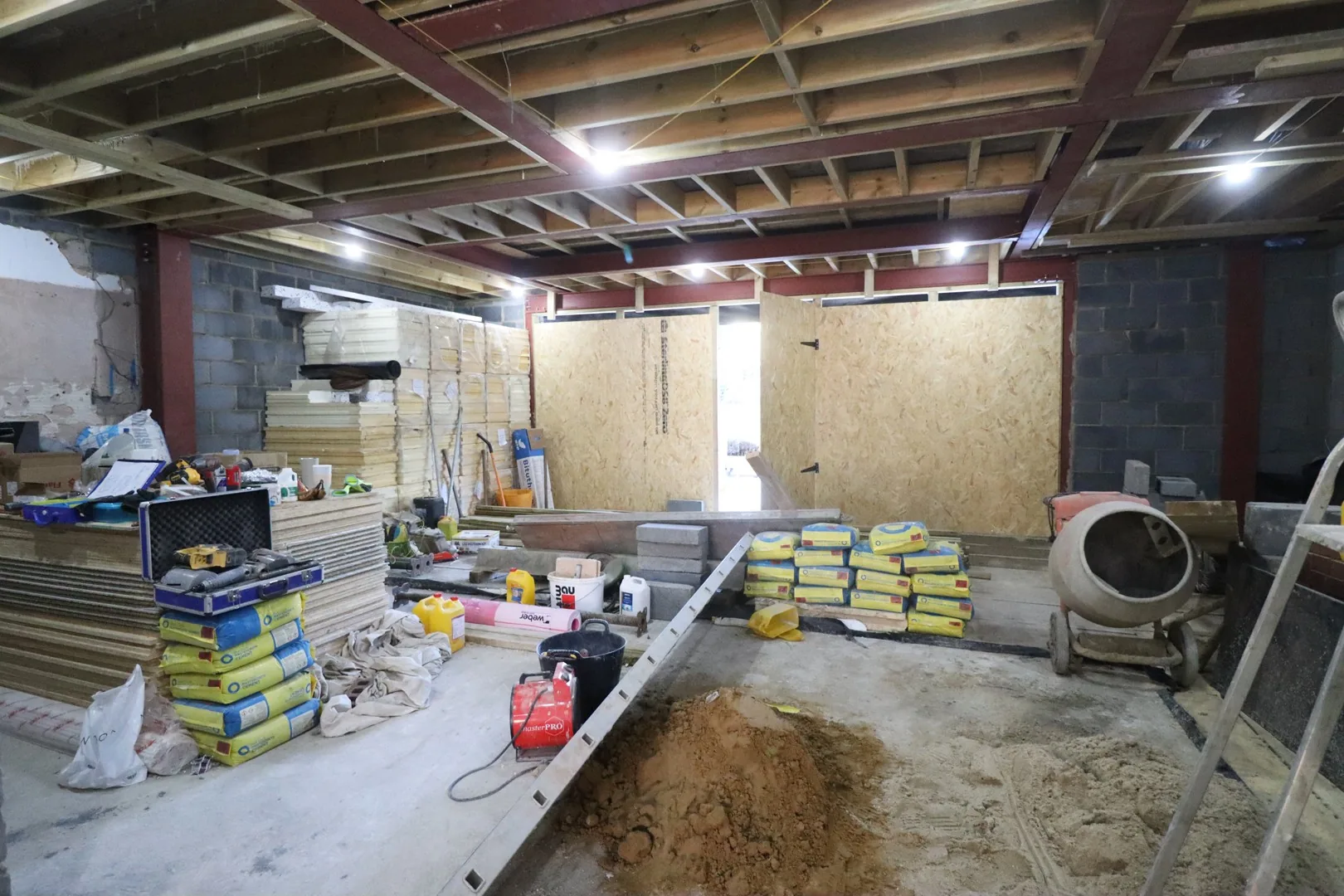 Ground floor interior during the rebuild: steel beams and new timber floor structure overhead, blockwork walls and boarded openings below.
