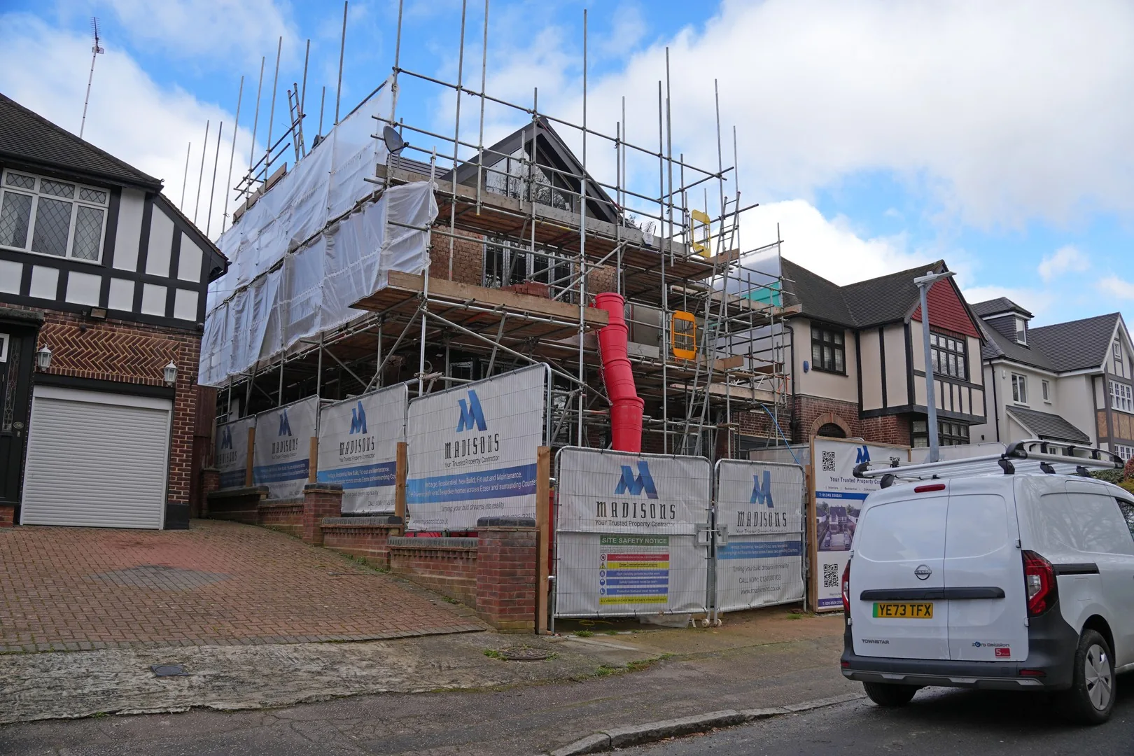 The Chigwell house from across the road, scaffolded between its neighbouring homes, with Madisons banners and the site safety notice on the fencing.
