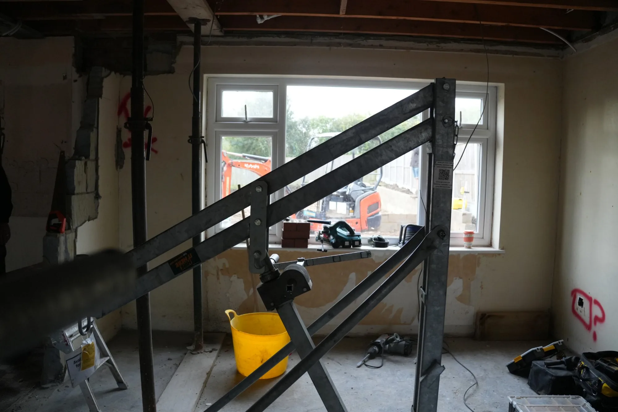 Props and a lifting hoist in place where a new structural opening is being formed, the digger at work through the window.