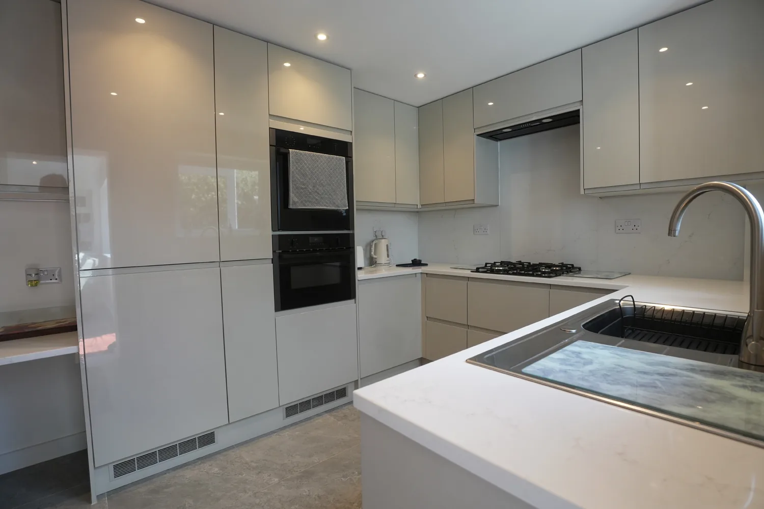 The new gloss handleless kitchen in the Witham house, with tall larder units, built-in double ovens and a white stone worktop with inset sink and swan neck tap.