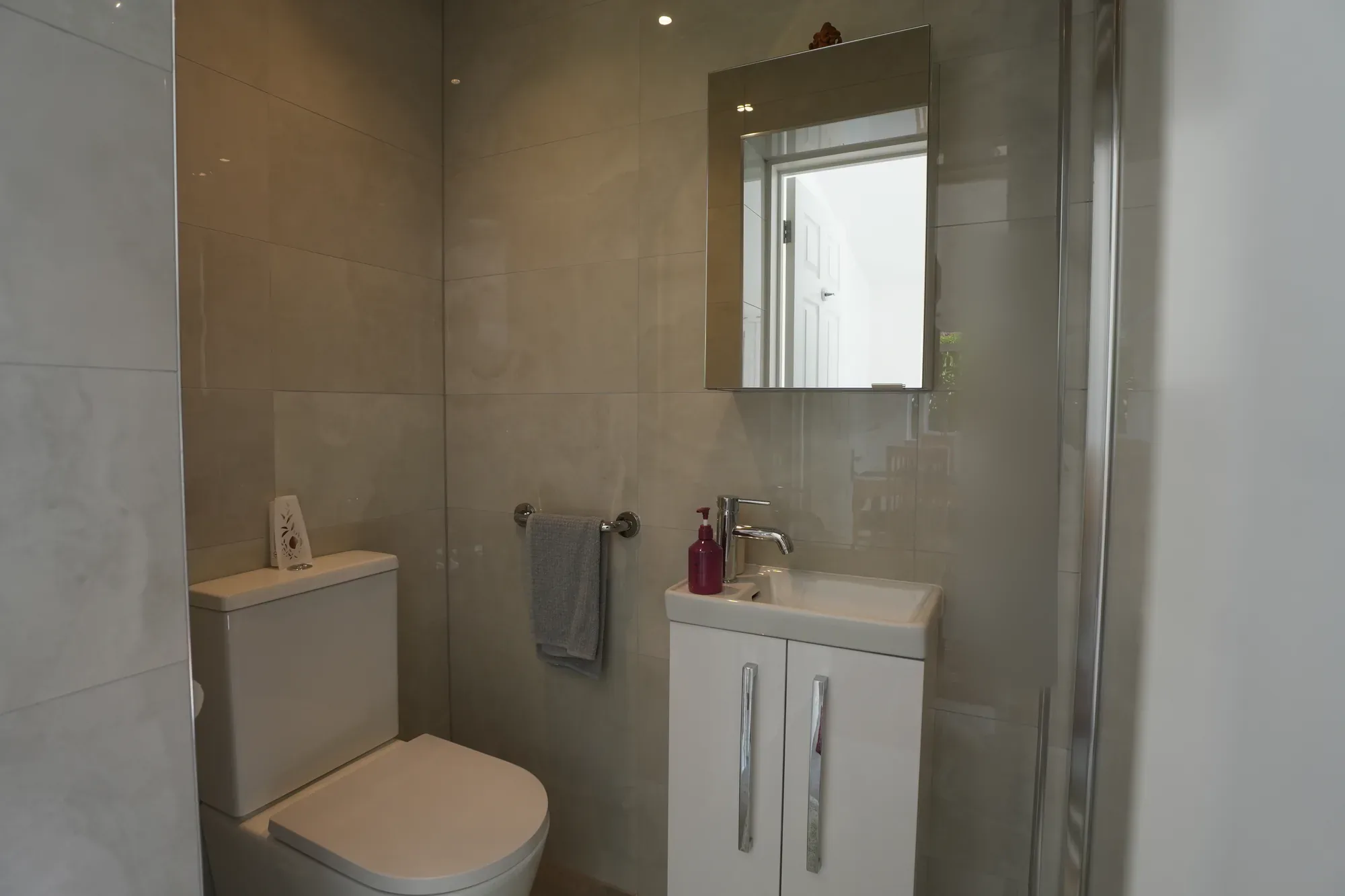 The Witham shower room basin and toilet beneath a wall mirror, with large format tiles and the edge of the glass shower screen.
